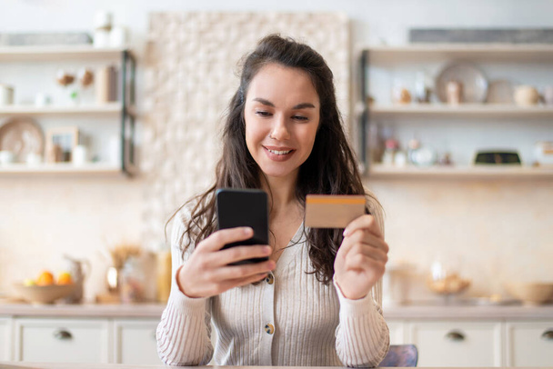 食料品ショッピングアプリ。クレジットカードや携帯電話を使用して陽気な女性,キッチンに座っている間にオンラインで食べ物を購入 - 写真・画像