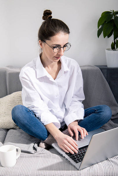 Giovane donna in occhiali con computer portatile con caffè seduto sul divano - Foto, immagini