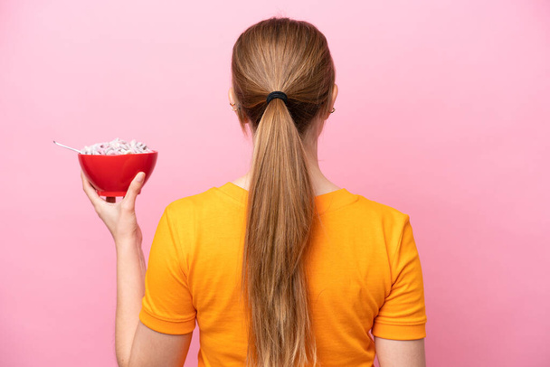 Young caucasian woman holding a bowl of cereals isolated on pink background in back position - Fotó, kép