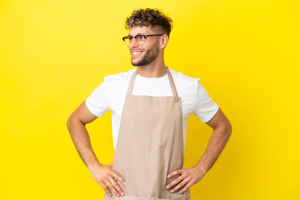 Restaurant waiter blonde man isolated on yellow background posing with arms at hip and smiling - Foto, afbeelding