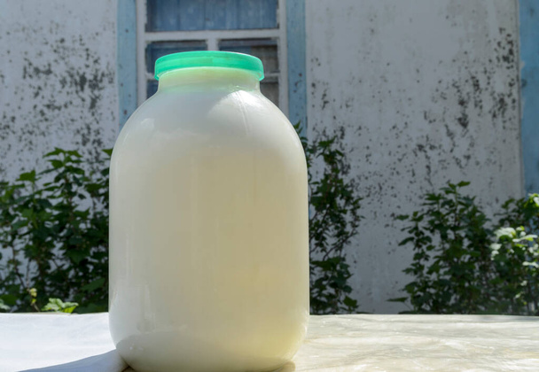 Frische Milch nach dem Melken auf einem Tisch in Glasflasche mit Deckel vor der Wand eines Dorfhauses mit Fenster und Büschen - Foto, Bild