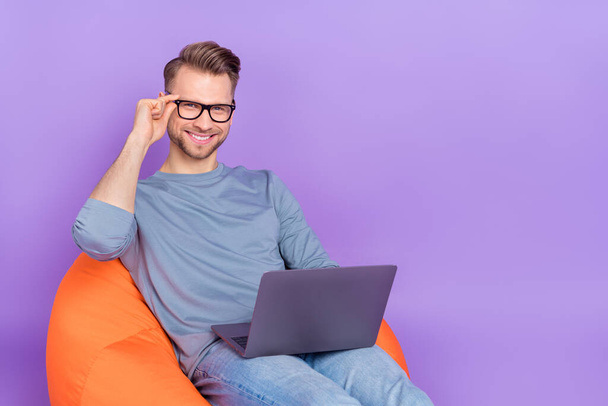 Photo of positive pretty guy wear grey shirt arm spectacles working device empty space sitting bean bag isolated violet color background - Foto, immagini