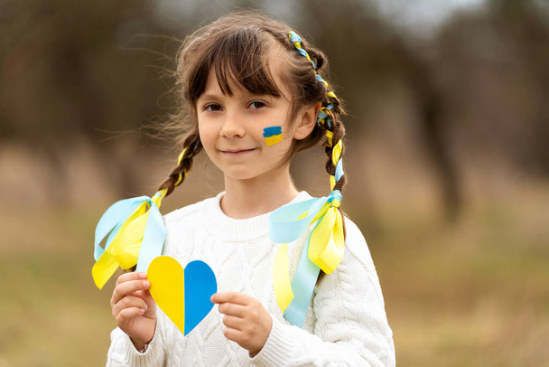 A little girl holds in her hands near her chest a heart in the patriotic colors of the Ukrainian flag, yellow and blue. Stop the war in Ukraine. Help save the Ukrainian nation. High quality photo - Φωτογραφία, εικόνα