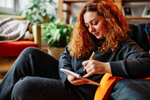 A cute young woman with red curly hair is studying and writing in her notebook while sitting in her living room. - Photo, image