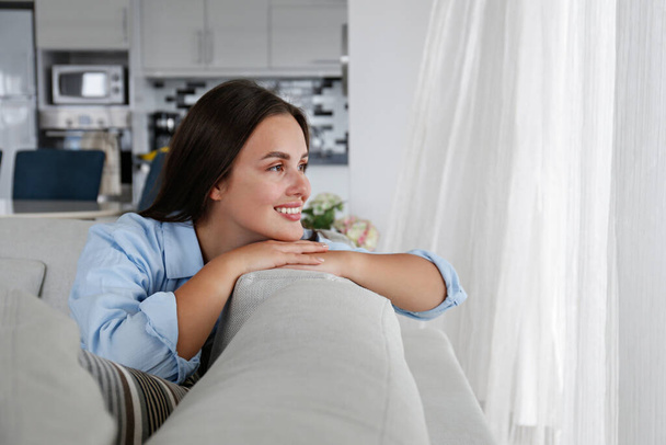 Portrait of young beautiful woman with long straight brunette hair sitting on the couch and smiling. Close up shot of brown eyed female with perfect smile. Copy space, background. - 写真・画像