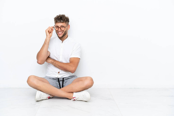 Young blonde man sitting on the floor isolated on white background with glasses and happy - Foto, Imagem