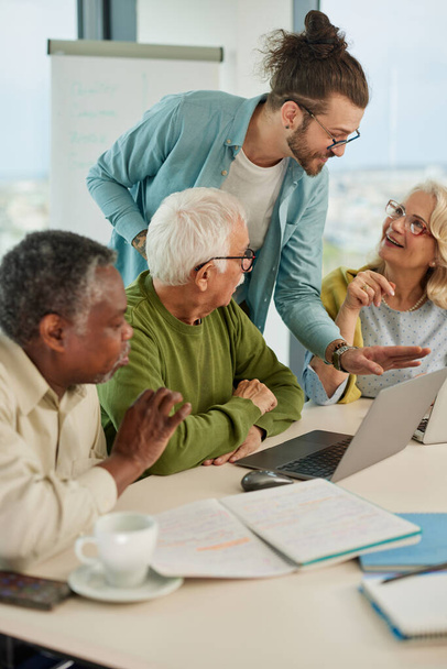 Un pequeño grupo de personas mayores multiculturales está teniendo una clase sobre las redes sociales en sus computadoras portátiles en el aula. - Foto, Imagen