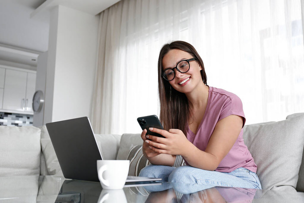 Portrait of young brunette woman wearing eyeglasses sitting at home, with the laptop scrolling the phone and smiling. Joyful female model having fun video call. Background, copy space, close up. - Valokuva, kuva