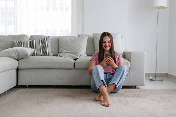 Portrait of young brunette woman wearing mom jeans sitting on the floor leaning on the couch texting and smiling. Joyful female having fun video phone call. Background, copy space, close up. - Fotografie, Obrázek