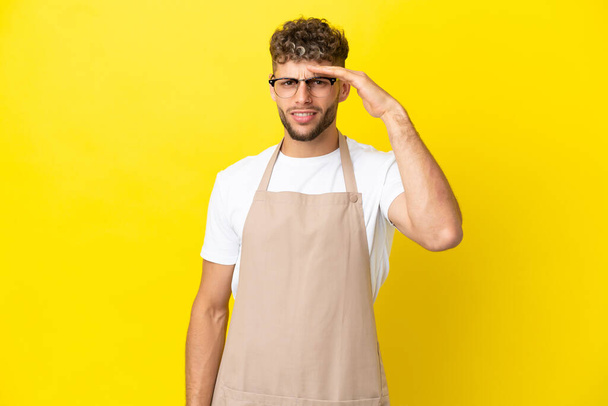 Restaurant waiter blonde man isolated on yellow background looking far away with hand to look something - Foto, imagen