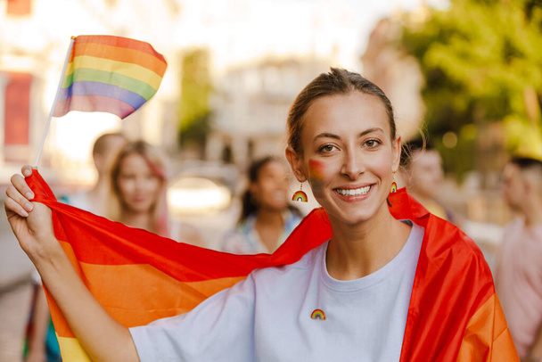 Young lesbian woman wrapped in rainbow flag smiling during pride parade on city street - Foto, afbeelding