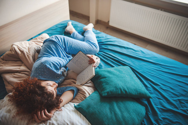 Кавказская кудрявая женщина удобно лежала на кровати, читая книгу. Кудрявая женщина читает книгу для дизайнеров модного образа жизни. - Фото, изображение
