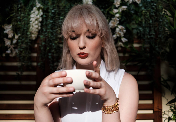 Πορτρέτο μιας νεαρής γυναίκας που πίνει καφέ. Φοράει λευκή μπλούζα και υπάρχουν φυτά πίσω της. Σάνι. - Φωτογραφία, εικόνα