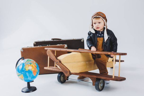 Poika nahkatakissa ja lentäjän hatussa, puinen lentokone, maapallo, ruskeat matkalaukut. Lasten puulelut - Valokuva, kuva
