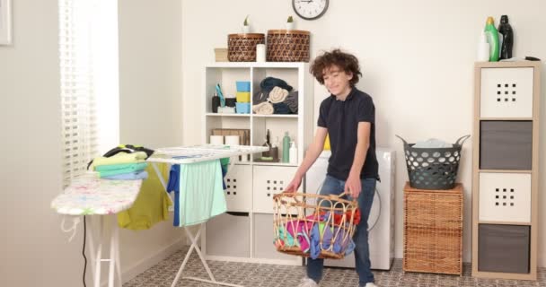 Jongen speelt met kleren tijdens de wasserij. jong tiener jongen gooien mand met wasgoed - Video