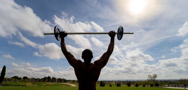 African man weightlifting outdoors,with his back against the light, sunny sky with clouds - Photo, Image