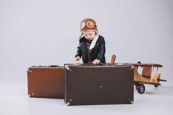 Šťastný chlapec v helmě a v lodivodské bundě nese hnědý kufr. Portrét dětského pilota, dítě v kožené bundě. Malý cestovatel s kufříkem - Fotografie, Obrázek