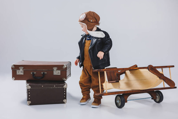 Um menino feliz em um capacete e uma jaqueta de piloto fica perto de um avião de madeira. Retrato de uma criança piloto, uma criança em uma jaqueta de couro. Brinquedos de madeira. Eco plano de árvore - Foto, Imagem
