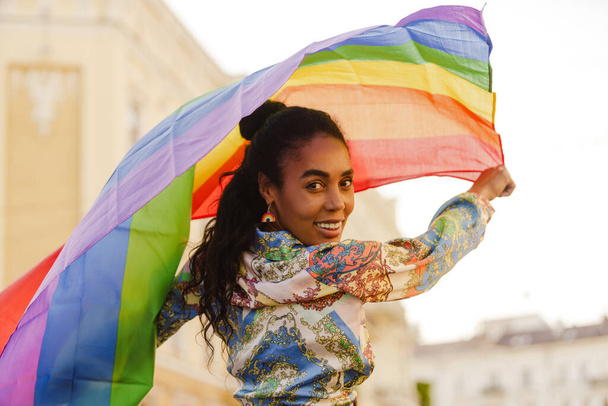Smiling african woman holding rainbow lgbt flag walking on a street - Photo, image