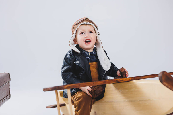 A happy boy in a helmet and a pilot's jacket stands near a wooden plane. Portrait of a child pilot, a child in a leather jacket. Wooden Toys. Eco plane from tree - 写真・画像