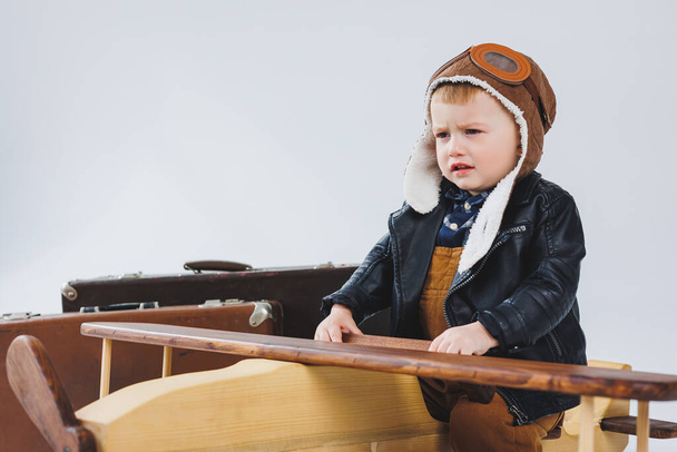 De jongen huilt in een leren jas en een pilotenhoed, een houten vliegtuig, bruine koffers. Houten kinderspeelgoed. Baby emoties - Foto, afbeelding