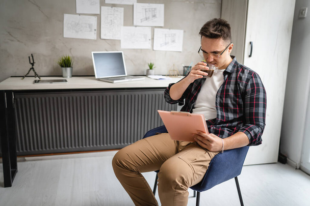 egy férfi fiatal kaukázusi férfi ül az íróasztal mellett a munkahelyen tanulmányi dokumentumok pihentető, miközben tartsa klip chart papírok valós emberek másolja tér férfi mérnök vagy diák élvezze a kávét - Fotó, kép