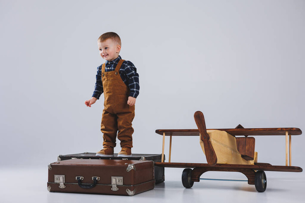 Un bambino allegro in una tuta marrone è in piedi su una valigia. Un piccolo viaggiatore con una valigia. Aereo in legno, giocattoli ecologici per bambini - Foto, immagini