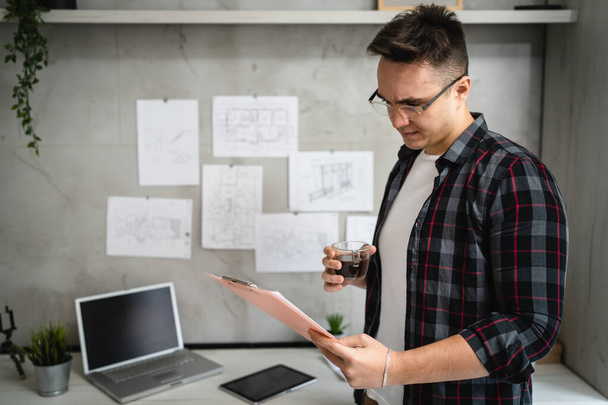 jeden mężczyzna młody kaukaski mężczyzna stojący przy biurku w pracy w biurze badania dokumentów relaks podczas trzymać klip wykres z papierami prawdziwi ludzie kopiować przestrzeń mężczyzna inżynier lub student cieszyć się kawa - Zdjęcie, obraz