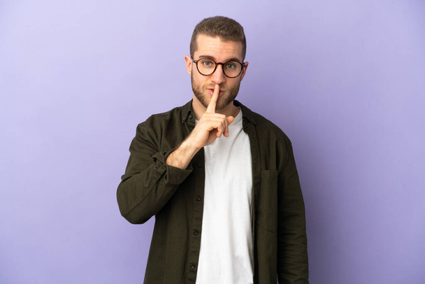 Young handsome caucasian man isolated on purple background showing a sign of silence gesture putting finger in mouth - Photo, Image
