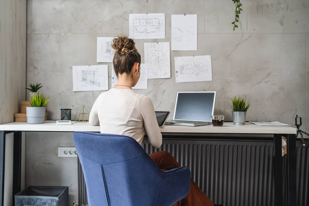 одна женщина, кавказка, сидящая за столом на работе в офисе, изучает документы, расслабляющие во время работы на ноутбуке; реальные люди копируют проект по планированию пространства женщины-инженера - Фото, изображение