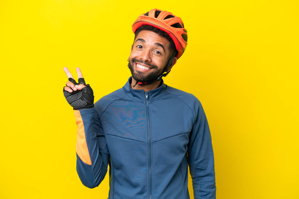 Junger brasilianischer Radrennfahrer auf gelbem Hintergrund lächelt und zeigt Siegeszeichen - Foto, Bild
