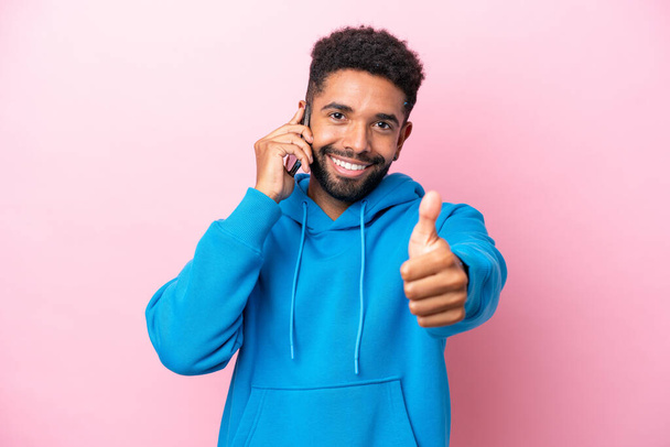 Young Brazilian man isolated on pink background keeping a conversation with the mobile while doing thumbs up - Фото, изображение