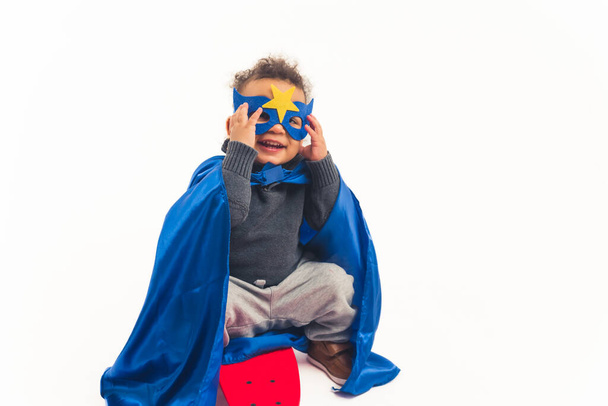 lindo afroamericano rizado pequeño niño con el traje de un superhéroe sentado en el monopatín sonriendo y tocando su máscara de estudio de tiro completo de fondo blanco espacio de copia - Foto, imagen