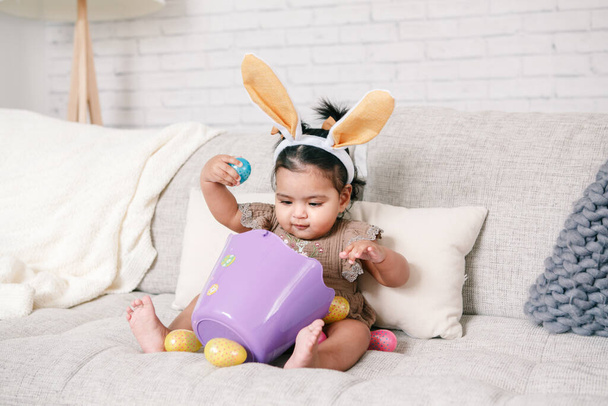 Cute Indian baby girl with pink bunny ears and basket of colorful eggs celebrating Easter holiday - Photo, Image