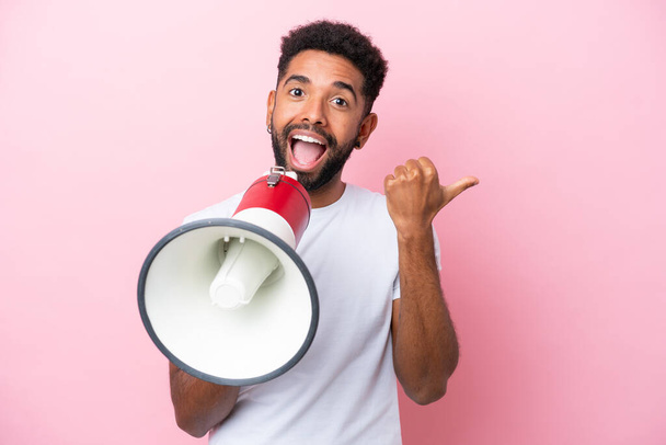 Young Brazilian man isolated on pink background shouting through a megaphone and pointing side - Foto, Imagem
