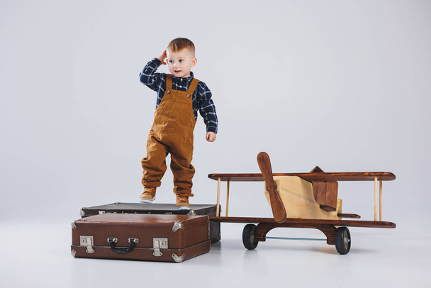 A cheerful child in a brown overalls is standing on a suitcase. Little traveler with a suitcase. Wooden plane, children's eco toys - 写真・画像