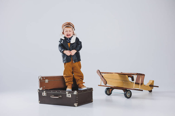 Il ragazzo piange con una giacca di pelle e un cappello da pilota, un aereo di legno, valigie marroni. Giocattoli in legno per bambini. Emozioni bambino - Foto, immagini