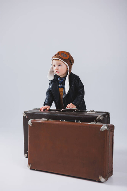 Um rapaz feliz com um capacete e um casaco de piloto carrega uma mala castanha. Retrato de uma criança piloto, uma criança em uma jaqueta de couro. Pequeno viajante com uma mala - Foto, Imagem