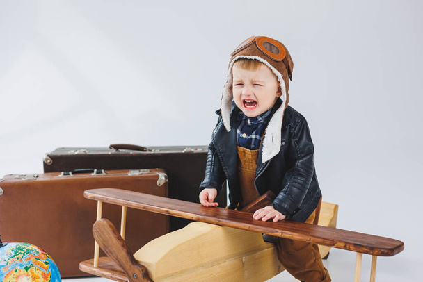El niño está llorando con una chaqueta de cuero y un sombrero de piloto, un avión de madera, maletas marrones. Juguetes de madera para niños. Emociones del bebé - Foto, Imagen