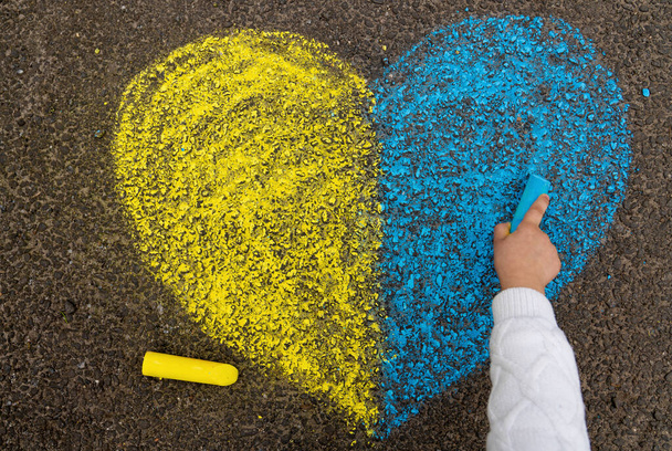 A child's hand draws a heart with crayons on the pavement with the patriotic national Ukrainian colors of yellow and blue. Stop the war in Ukraine. High quality photo - 写真・画像