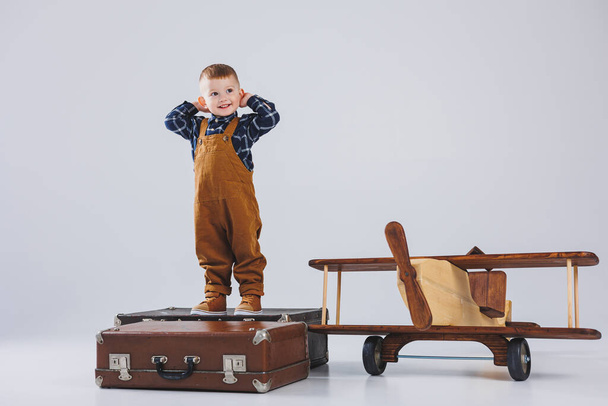 A cheerful child in a brown overalls is standing on a suitcase. Little traveler with a suitcase. Wooden plane, children's eco toys - Photo, Image