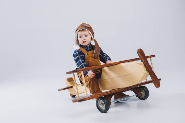 Kleine jongen van 3 jaar oud speelt met een houten vliegtuig op een witte achtergrond. In een overall en een pilotenhoed. Kinder eco speelgoed gemaakt van natuurlijk hout. houten vliegtuig - Foto, afbeelding