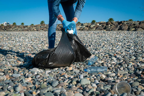 Woman collects plastic, foam, rubber in a black garbage bag on a pebble beach on a sunny day. Cleaning of the coastal zone. The concept of ecology, volunteering - Photo, Image