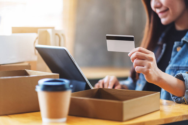 Nahaufnahme einer jungen asiatischen Frau mit Tablet-PC und Kreditkarte beim Online-Einkauf mit Postpaketkasten und Einkaufstaschen auf dem Tisch - Foto, Bild