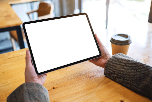Immagine mockup di una donna che tiene in mano tablet pc nero con schermo bianco vuoto con tazza di caffè su tavolo di legno - Foto, immagini
