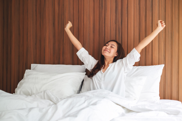 Egy gyönyörű ázsiai nő nyújtózkodik, miután reggel felébredt egy fehér, kényelmes ágyon otthon. - Fotó, kép