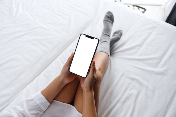 Imagem de maquete vista superior de uma mulher segurando telefone celular branco com tela de desktop em branco enquanto deitado em uma cama branca aconchegante em casa - Foto, Imagem