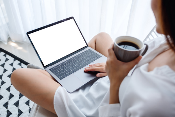 自宅のベッドルームでコーヒーを飲みながら、空白の画面を持つラップトップコンピュータで作業して入力する女性のモックアップイメージ - 写真・画像