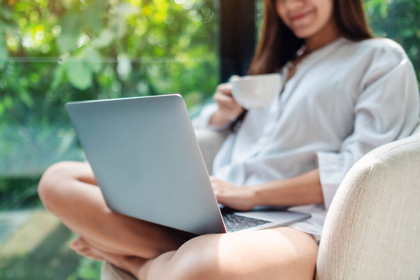 Close-up beeld van een vrouw die werkt op laptop computer tijdens het drinken van koffie en zitten op een witte fauteuil thuis met groene natuur achtergrond - Foto, afbeelding