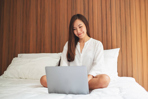 Szoros kép egy gyönyörű ázsiai nő használja és dolgozik a laptop számítógép, miközben ül egy fehér kényelmes ágy otthon - Fotó, kép